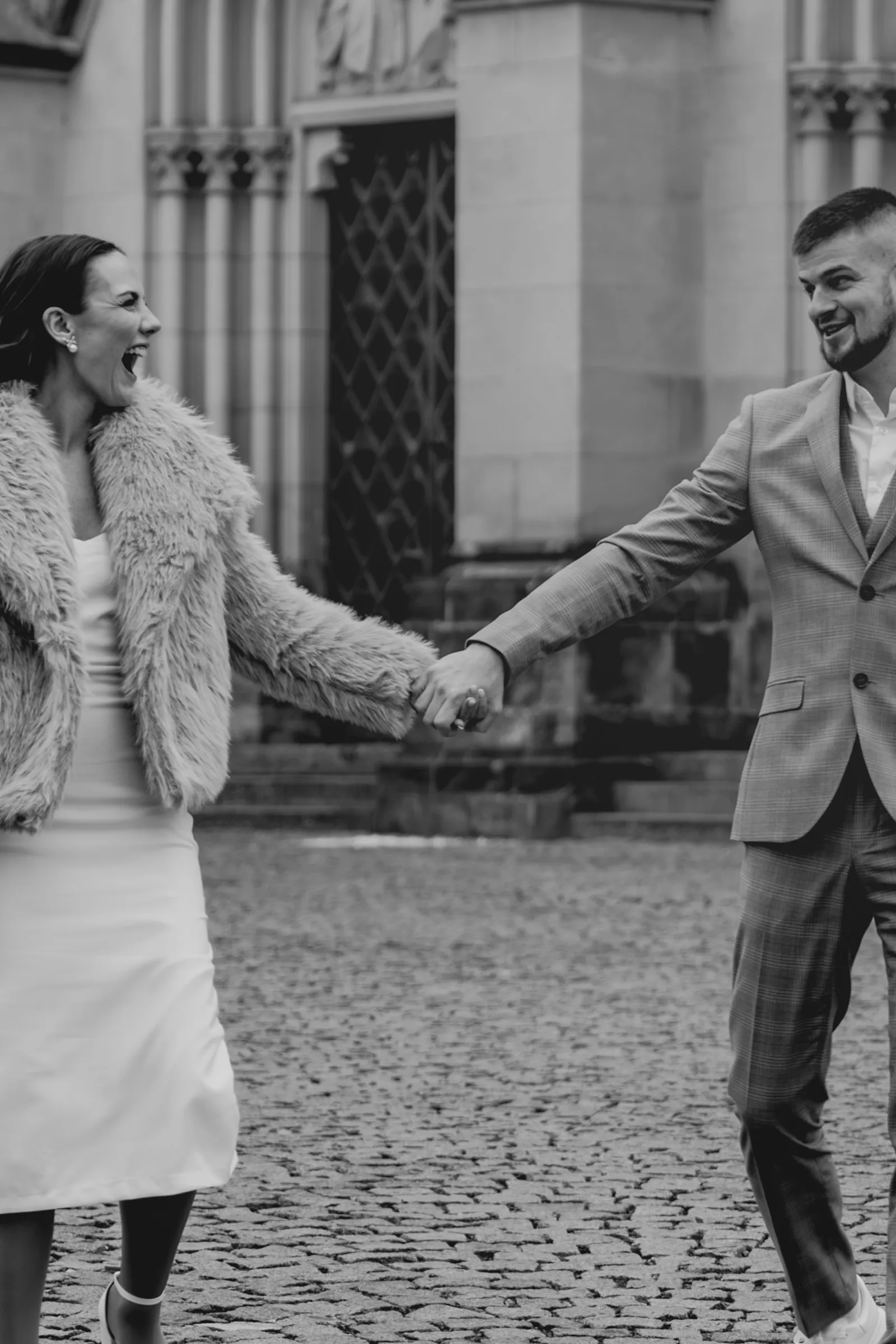 Černobílý záběr rozesmáté nevěsty v kožíšku a ženicha, kteří se drží za ruce na dlážděném náměstí
