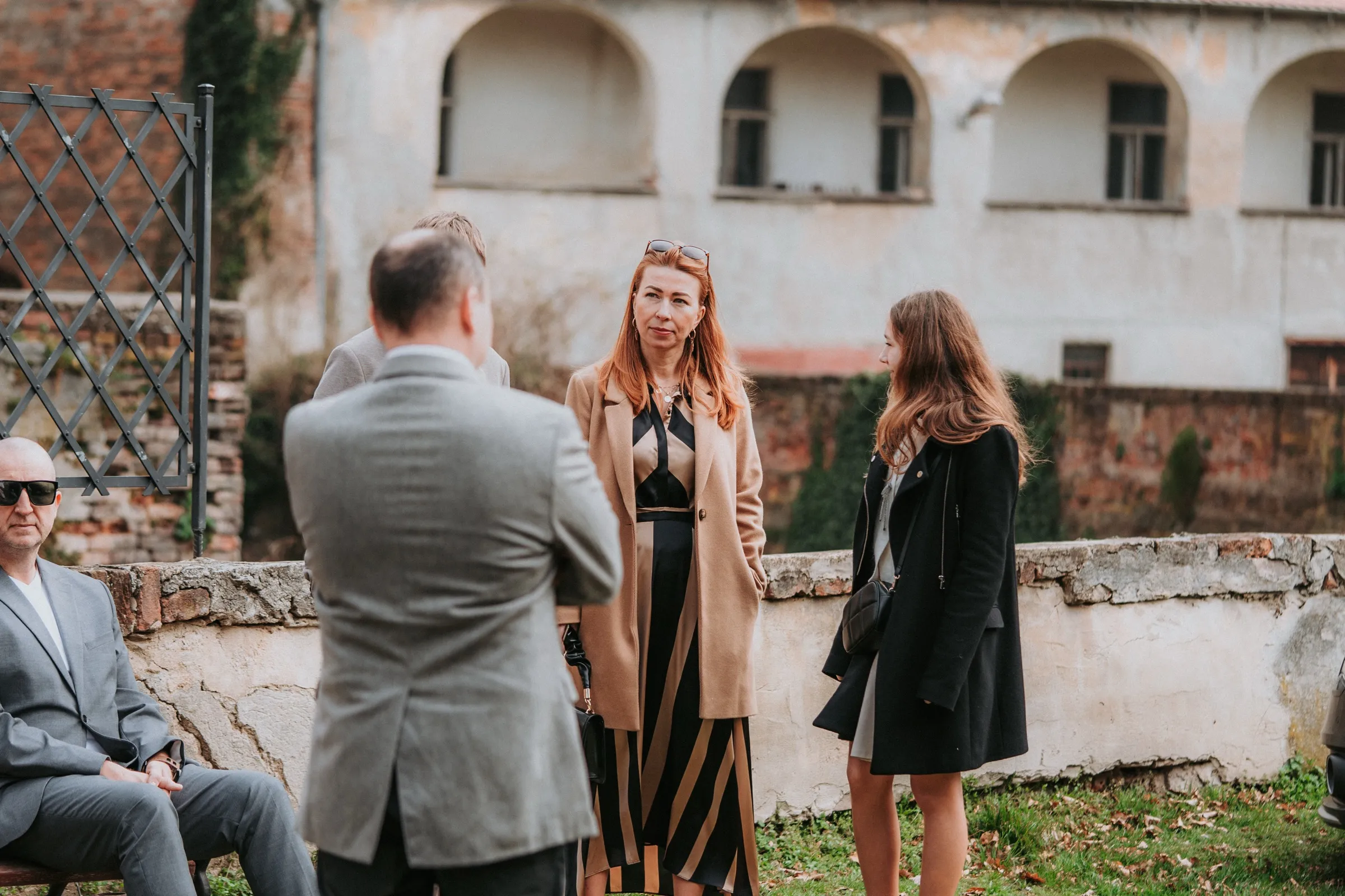 Svatební hosté v kabátech čekají na zahradě u staré zídky, v pozadí budova s arkádami