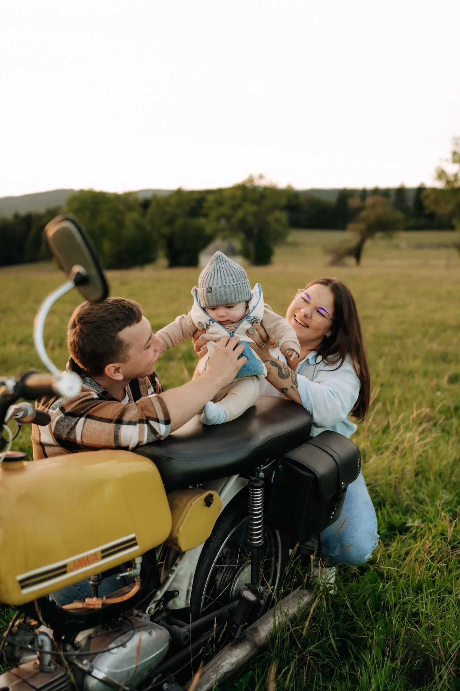Rodiče si na louce hrají s miminkem posazeným na žlutém motocyklu